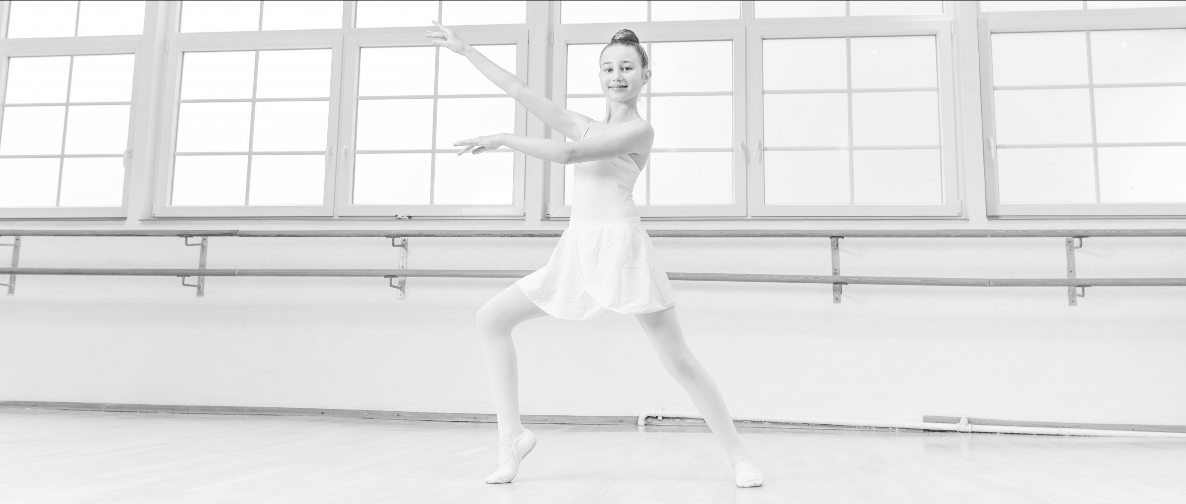 Junge Ballerina im weißen Ballettoutfit steht in der Tanzschule & Ballettschule vor der Stange in eleganter Pose